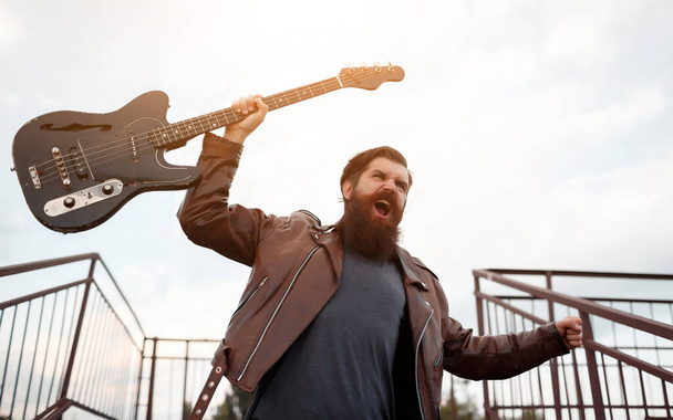 Un brutal rockero barbudo con pelo gris en una chaqueta de cuero marrón y vaqueros azules sostiene una guitarra eléctrica en su mano y grita contra el fondo de una escalera peatonal que cruza - Foto, imagen