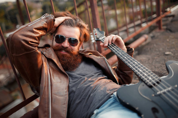 Бородатий чоловік з довгими вусами в чорних окулярах і коричнева шкіряна куртка лежить тримаючи чорну гітару і сміється
 - Фото, зображення