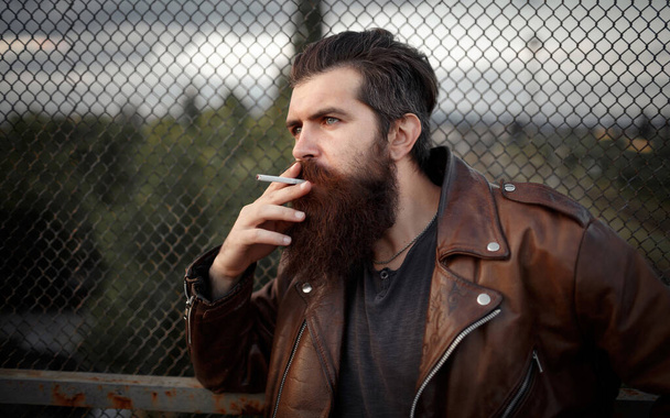 Męski rocker z długą brodą, wąsami i siwymi włosami w brązowej skórzanej kurtce pali papierosy i patrzy w dal na tle żelaznej siatki - Zdjęcie, obraz