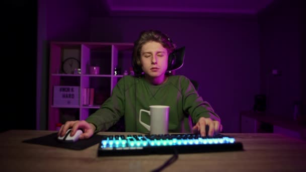 Vermoeide jongeman zittend in een speelstoel in de kamer met een paars licht, videospelletjes spelend op de computer en koffie drinkend uit een kopje. Nachtspelen. - Video