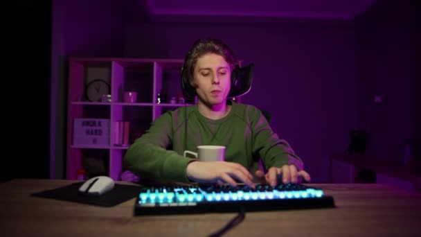 Retrato de un jugador cansado en un auricular con una taza en sus manos jugando juegos en línea en el ordenador y mirando atentamente a la cámara. - Imágenes, Vídeo