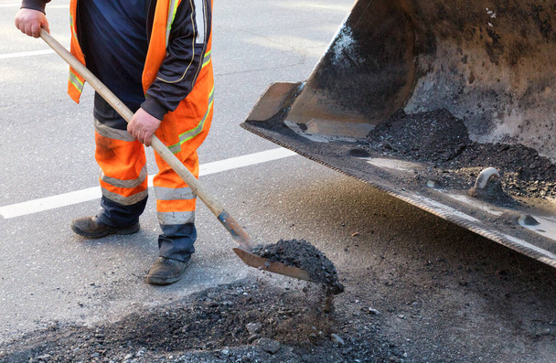 Un operaio del servizio stradale infila un vecchio asfalto nella benna metallica di un escavatore mentre ripara un tratto della strada. - Foto, immagini