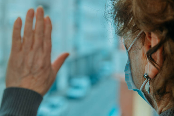 Yüz maskesi takmış yaşlı bir kadın, elleri penceresinde eve kapatılmış. - Fotoğraf, Görsel