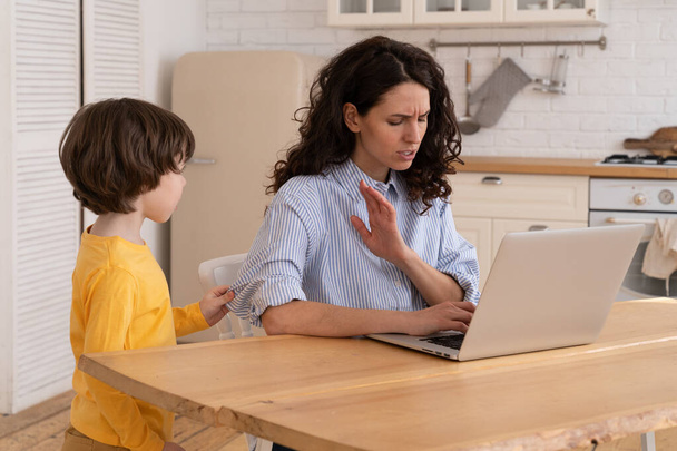 Мама сидит за столом в домашнем офисе во время блокировки, работает на ноутбуке, ребенок отвлекает и шуметь - Фото, изображение