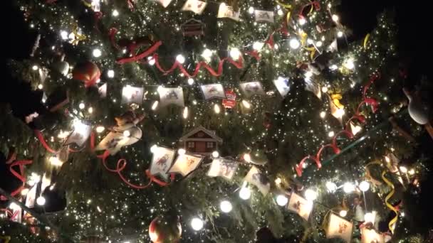 geschmückter großer Weihnachtsbaum mit Hintergrundbeleuchtung auf der Straße. Flaggen Spielzeug und Glühbirnen - Filmmaterial, Video