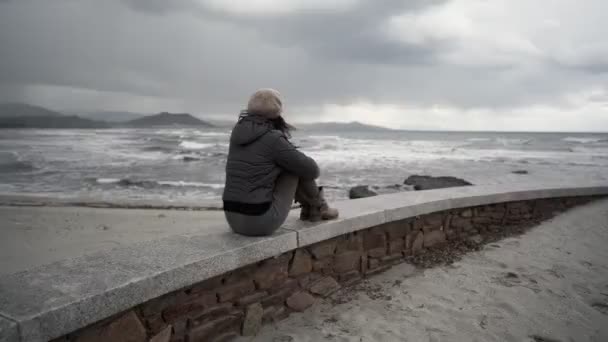 Onherkenbare jonge vrouw in de winter jurk en wollen hoed zittend op een muur door de kust schot van achteren kijken naar de stormachtige oceaan golven onder een dramatische bewolkte lucht. Pensioen persoon blik horizon - Video