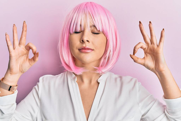 Jovem loira vestindo peruca rosa relaxar e sorrir com os olhos fechados fazendo gesto de meditação com os dedos. conceito de ioga.  - Foto, Imagem