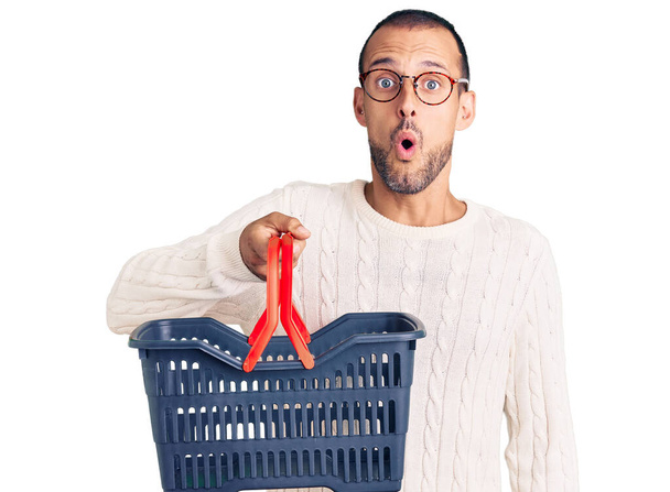 Young handsome man holding supermarket shopping basket scared and amazed with open mouth for surprise, disbelief face  - 写真・画像