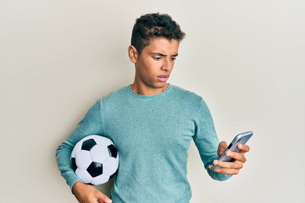 Fiatal jóképű afro-amerikai férfi kezében focilabda nézi smartphone sokk arc, látszó szkeptikus és szarkasztikus, meglepett nyitott szájjal  - Fotó, kép