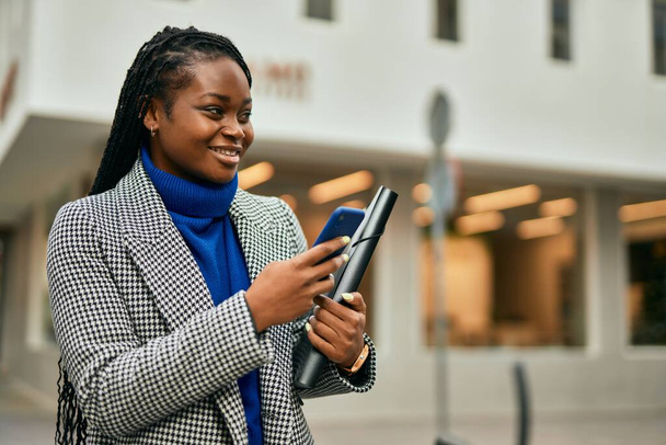 Νεαρή Αφροαμερικανή επιχειρηματίας χαμογελώντας ευτυχισμένη χρησιμοποιώντας smartphone στην πόλη. - Φωτογραφία, εικόνα