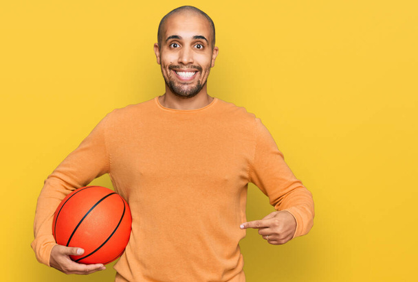 Hispanic adult man holding basketball ball smiling happy pointing with hand and finger  - Фото, изображение