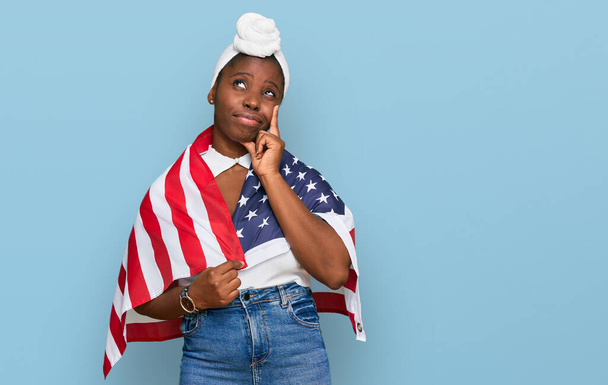 Молодая африканская женщина с тюрбаном, держащая флаг Соединенных Штатов серьезное лицо, думая о вопросе с рукой на подбородке, вдумчивый о запутанной идее  - Фото, изображение