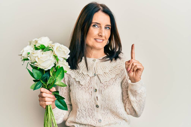 Μέση ηλικία μελαχρινή γυναίκα κρατώντας μπουκέτο από λευκά λουλούδια χαμογελώντας με μια ιδέα ή ερώτηση δείχνοντας δάχτυλο με χαρούμενο πρόσωπο, νούμερο ένα  - Φωτογραφία, εικόνα