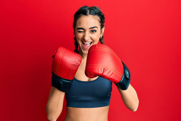 Young brunette girl using boxing gloves winking looking at the camera with sexy expression, cheerful and happy face.  - Photo, Image