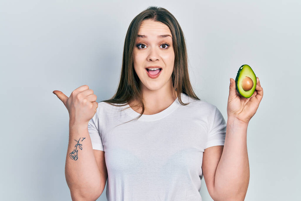 jong hispanic meisje houden midden avocado wijzen duim naar de zijkant glimlachen blij met open mond  - Foto, afbeelding