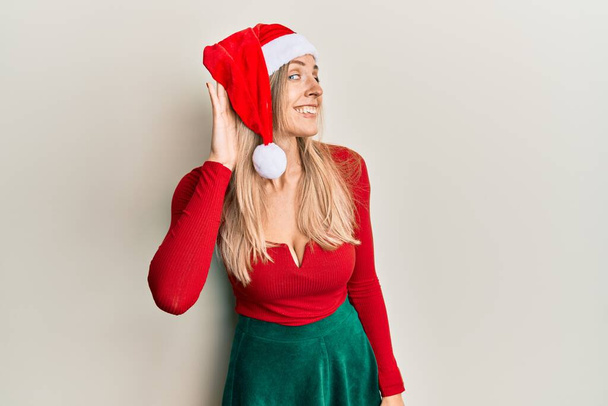 Mulher caucasiana bonita vestindo traje de Natal e chapéu sorrindo com a mão sobre a orelha ouvindo uma audição para boatos ou fofocas. conceito de surdez.  - Foto, Imagem