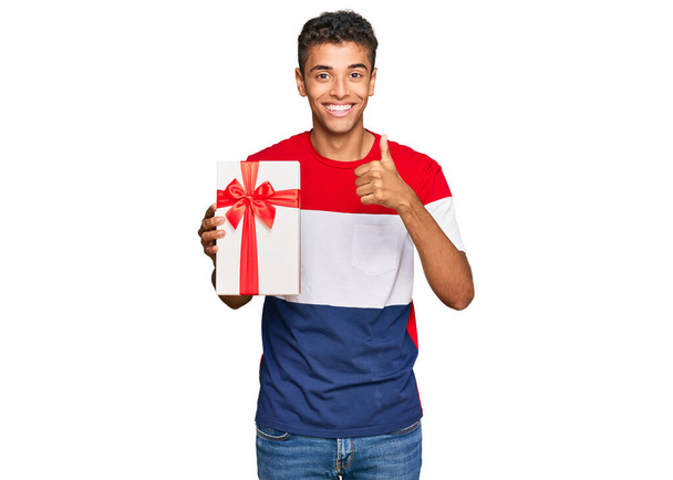 Młody przystojny Afroamerykanin mężczyzna trzyma prezent uśmiechnięty szczęśliwy i pozytywny, kciuk do góry robi doskonałe i znak aprobaty  - Zdjęcie, obraz