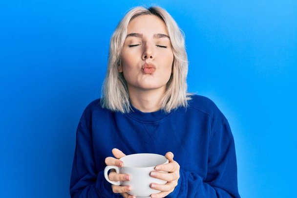 Mladá blondýnka pije šálek kávy, dívá se do kamery a posílá polibek, který je krásný a sexy. výraz lásky.  - Fotografie, Obrázek