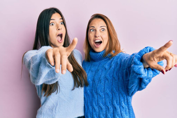 Hispanic family of mother and daughter wearing wool winter sweater pointing with finger surprised ahead, open mouth amazed expression, something on the front  - Fotoğraf, Görsel