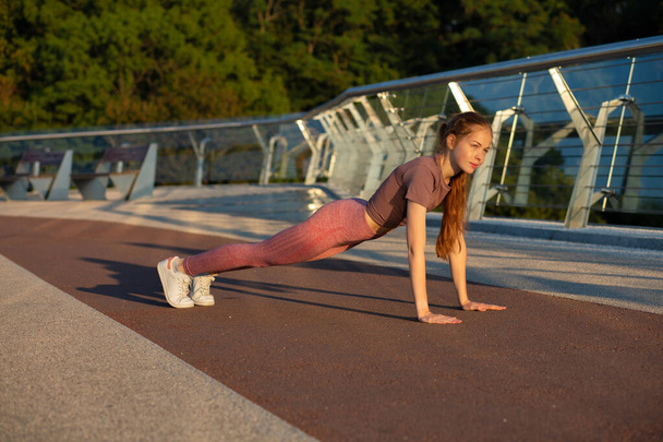 Athletic red head fitness woman in sport apparel doing plank exercise at the bridge - Photo, Image