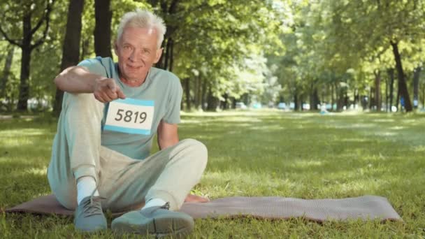 Szerokie ujęcie dobrze wyglądającego białego starszego sportowca siedzącego na gumowej macie w parku, patrzącego w kamerę, wskazującego palcem na ekran i zapraszającego na trening na świeżym powietrzu - Materiał filmowy, wideo