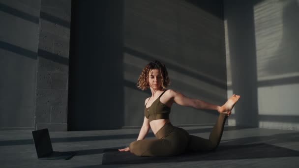 Mujer atractiva joven practicando ejercicios de estiramiento gimnástico usando su computadora. Concepto de formación de yoga online - Imágenes, Vídeo