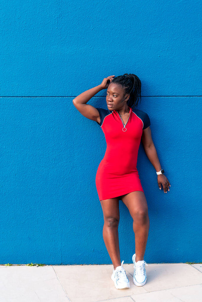 negro Afro chica posando en la calle sobre un fondo azul en un colorido vestido rojo - Foto, imagen