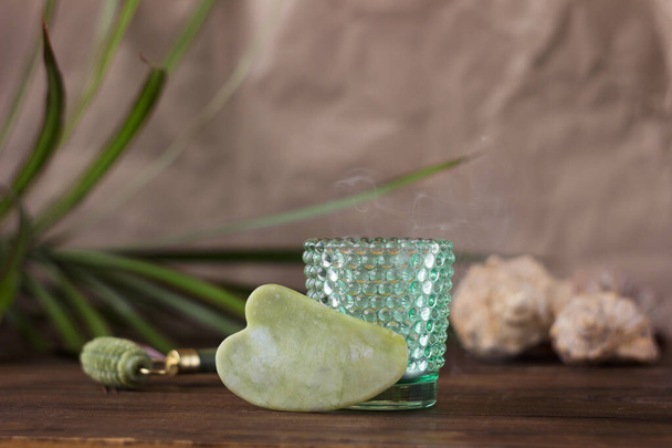 Set de masajes: gouache verde y rodillo facial de jade. Fondo de madera, hojas de plantas, vela en un vidrio con espinillas - Foto, Imagen