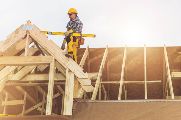 Entrepreneur caucasien avec le niveau d'esprit dans sa main sur le dessus de la structure de toit de maison en bois nouvellement construite. Thème industriel. - Photo, image