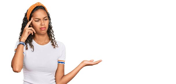 カジュアルな服を着ている若いアフリカ系アメリカ人の少女は、コピースペースと額に指を指すことを示すオープン手のひらに混乱し、悩まされています。考えてみてください.  - 写真・画像