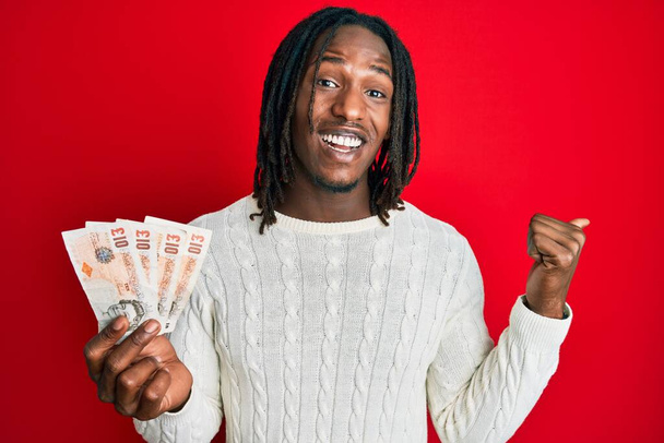 African american man with braids holding 10 united kingdom pounds banknotes pointing thumb up to the side smiling happy with open mouth  - Foto, Bild