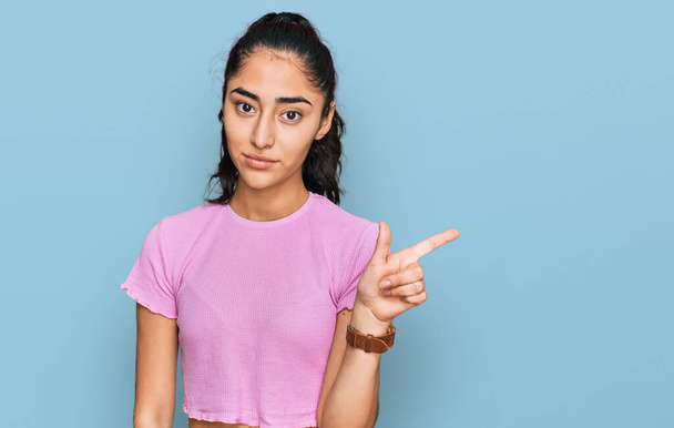 Латиноамериканская девочка-подросток с зубными скобками в повседневной одежде, указывая пальцем в сторону, показывая рекламу, серьезное и спокойное лицо  - Фото, изображение