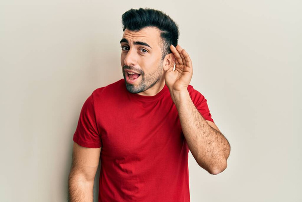 Jonge Spaanse man in casual kleding glimlachend met hand over oor luisterend en horend naar geruchten of roddels. doofheid.  - Foto, afbeelding