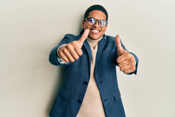 エレガントな服と眼鏡を身に着けている若いハンサムなヒスパニック系の男性は、手で積極的なジェスチャーを行うことを承認し、親指を笑顔と成功のために幸せ。勝者のジェスチャー.  - 写真・画像