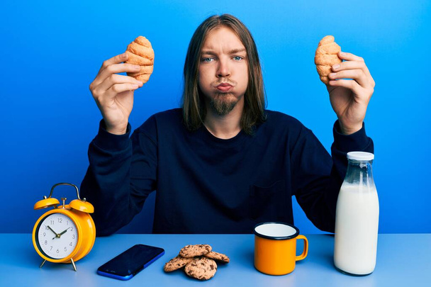 Komea valkoihoinen mies, jolla on pitkät hiukset ja joka syö croissantia aamiaiseksi. ilmatäytteinen suu, raikas ilma.  - Valokuva, kuva