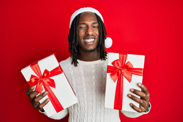 アフリカ系アメリカ人男性とともにbrids身に着けているクリスマスの帽子と保持ギフトwinking見ますザカメラとともにセクシー表現,陽気で幸せな顔.  - 写真・画像