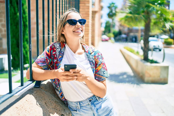 Joven hermosa rubia caucásica mujer sonriendo feliz al aire libre en un día soleado con el teléfono inteligente - Foto, imagen