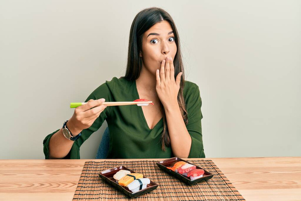 Νεαρή μελαχρινή γυναίκα που τρώει σούσι κάθεται στο τραπέζι καλύπτοντας το στόμα με το χέρι, σοκαρισμένη και φοβισμένη για λάθος. έκπληκτη έκφραση  - Φωτογραφία, εικόνα