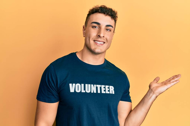 Spanischer junger Mann im freiwilligen T-Shirt lächelt fröhlich und zeigt mit der Handfläche in die Kamera.  - Foto, Bild