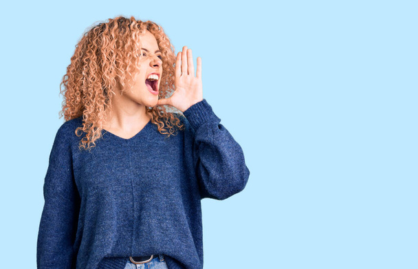 Junge blonde Frau mit lockigem Haar trägt lässigen Winterpullover und schreit laut Seite an Seite mit der Hand auf dem Mund. Kommunikationskonzept.  - Foto, Bild