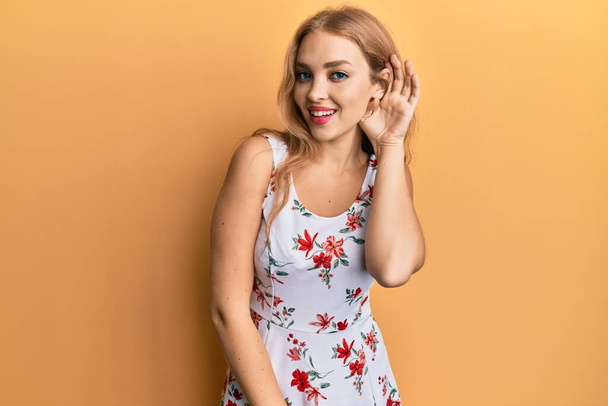 Beautiful blonde caucasian woman wearing floral dress smiling with hand over ear listening and hearing to rumor or gossip. deafness concept.  - Fotografie, Obrázek