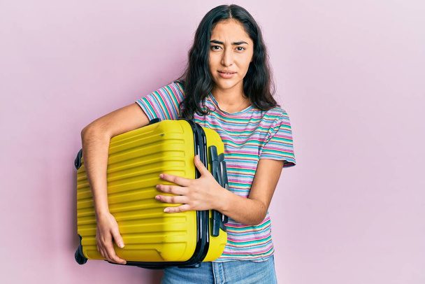 Hispanic teenager girl with dental braces holding cabin bag clueless and confused expression. doubt concept.  - Φωτογραφία, εικόνα