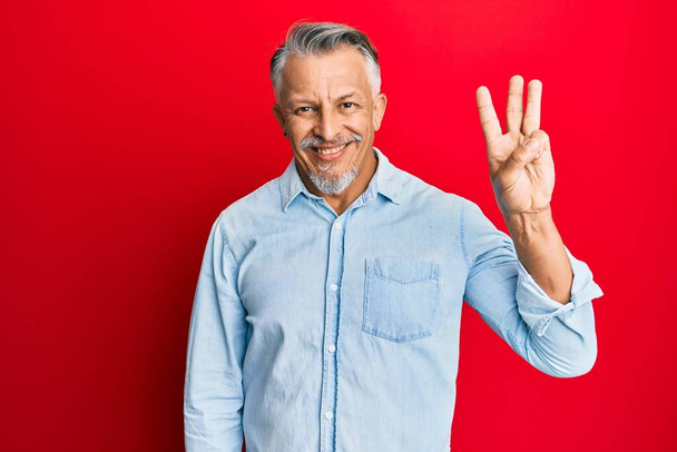 Grauhaariger Mann mittleren Alters in lässiger Kleidung, der mit Finger Nummer drei zeigt und nach oben zeigt, während er selbstbewusst und glücklich lächelt.  - Foto, Bild
