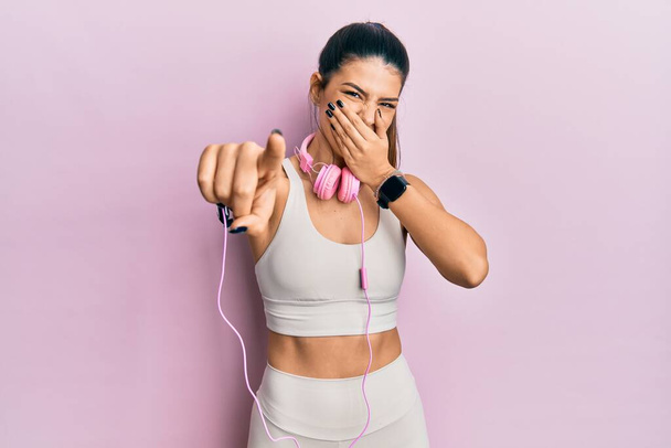 Mujer hispana joven usando ropa de gimnasia y usando auriculares riéndose de ti, señalando con el dedo a la cámara con la boca sobre la mano, expresión de vergüenza  - Foto, Imagen