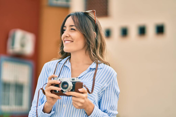 Młoda latynoska turystka uśmiechnięta szczęśliwa używając zabytkowej kamery w mieście. - Zdjęcie, obraz