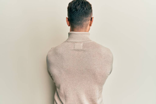 Hispanic young man wearing casual turtleneck sweater standing backwards looking away with crossed arms  - Fotó, kép