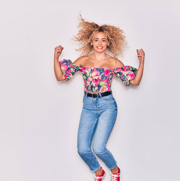 Young beautiful curly girl wearing casual clothes smiling happy. Jumping with smile on face celebrating with fists up over isolated white background. - Photo, Image