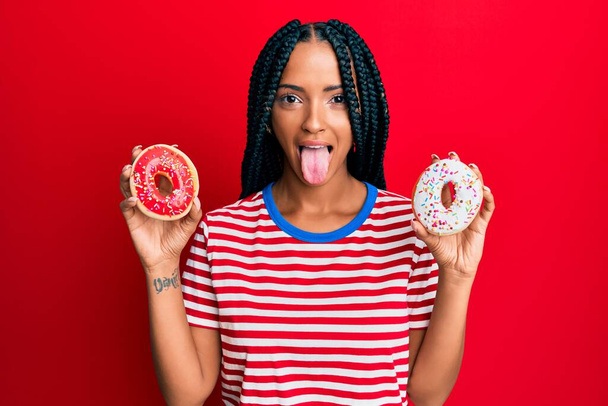 Beautiful hispanic woman holding tasty colorful doughnuts sticking tongue out happy with funny expression.  - Foto, imagen