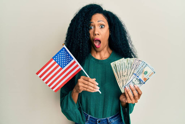 Középkorú afro-amerikai nő kezében Egyesült Államok zászló és dollár fél, és megdöbbent a meglepetés és csodálkozó kifejezés, félelem és izgatott arc.  - Fotó, kép