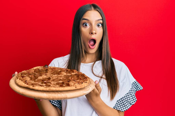 Молодая латиноамериканка, держащая итальянскую пиццу в страхе и шоке от удивления и удивления, страха и возбужденного лица.  - Фото, изображение